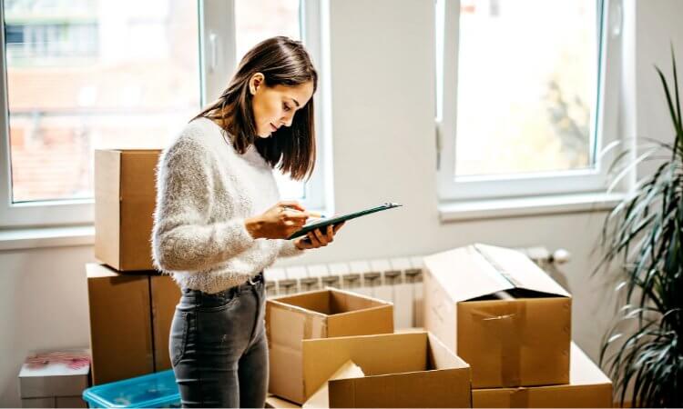 Find moving helpers in your area Young woman standing between moving boxes and looking at her smartphone, symbolising simple organisation and proven assistance with moving with KnowS.